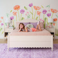 Young girl lying on pink bed in pastel floral girls' bedroom with large flower wall decals, bedside lamps, lavender rug and soft bedding — kids room decor and tween bedroom ideas