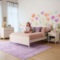 Smiling young girl sitting on a pink bed in a pastel girls' bedroom with watercolor floral wall decals, white nightstand, pastel lamp, purple shag rug and cozy bedding — kids room decor inspiration