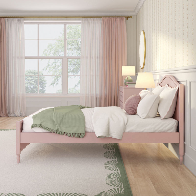 Pastel pink bedroom with pink bed frame, green throw blanket, layered sheer curtains, bedside lamp, round mirror and subtle patterned wallpaper