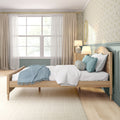 Sunlit cozy bedroom interior with wooden bed, teal pillows and throw, floral wallpaper, sheer curtains over a large window and bedside lamp