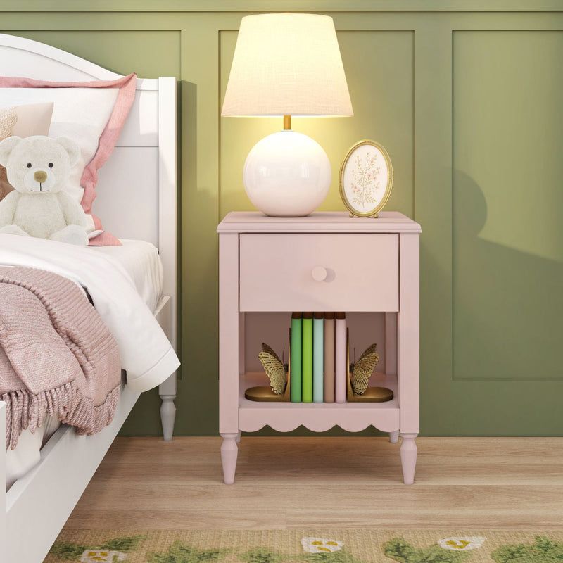 Pink bedside table with drawer and shelf holding pastel books and butterfly bookends, topped by a white globe lamp and framed floral art beside a teddy bear on a white bed against a green paneled accent wall