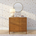 Mid-century wooden dresser with cream ceramic lamp and round brass mirror on cream floral wallpaper, pink headphones on books, cozy neutral bedroom decor
