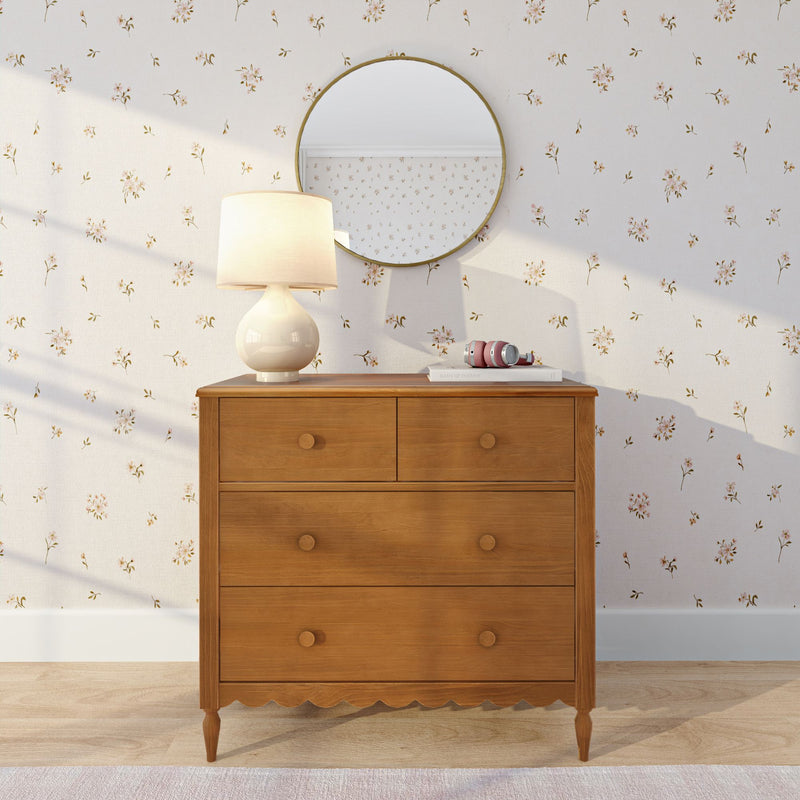 Mid-century wooden dresser with cream ceramic lamp and round brass mirror on cream floral wallpaper, pink headphones on books, cozy neutral bedroom decor