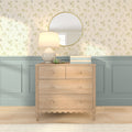 Wooden dresser with lamp, round gold mirror, stacked books and headphones against floral wallpaper and pastel blue wainscoting