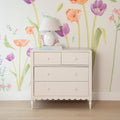 Light pink wooden dresser topped with a white ceramic table lamp and books against pastel watercolor tulip and poppy wallpaper