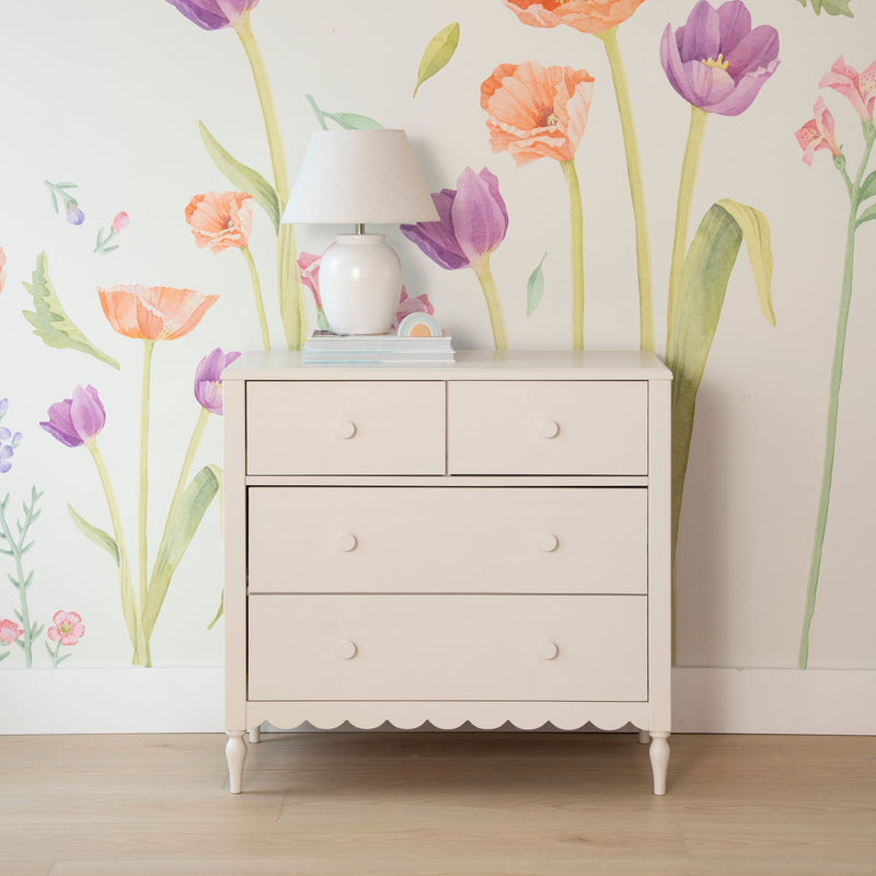 Light pink wooden dresser topped with a white ceramic table lamp and books against pastel watercolor tulip and poppy wallpaper