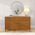 Wooden dresser with round gold wall mirror reflecting a potted plant, cream table lamp and pink headphones on floral wallpaper, styled bedroom dresser decor
