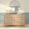 Light wood dresser topped with a ceramic table lamp, stacked books and a round gold mirror reflecting a window, set against cream floral wallpaper and pastel blue wainscoting