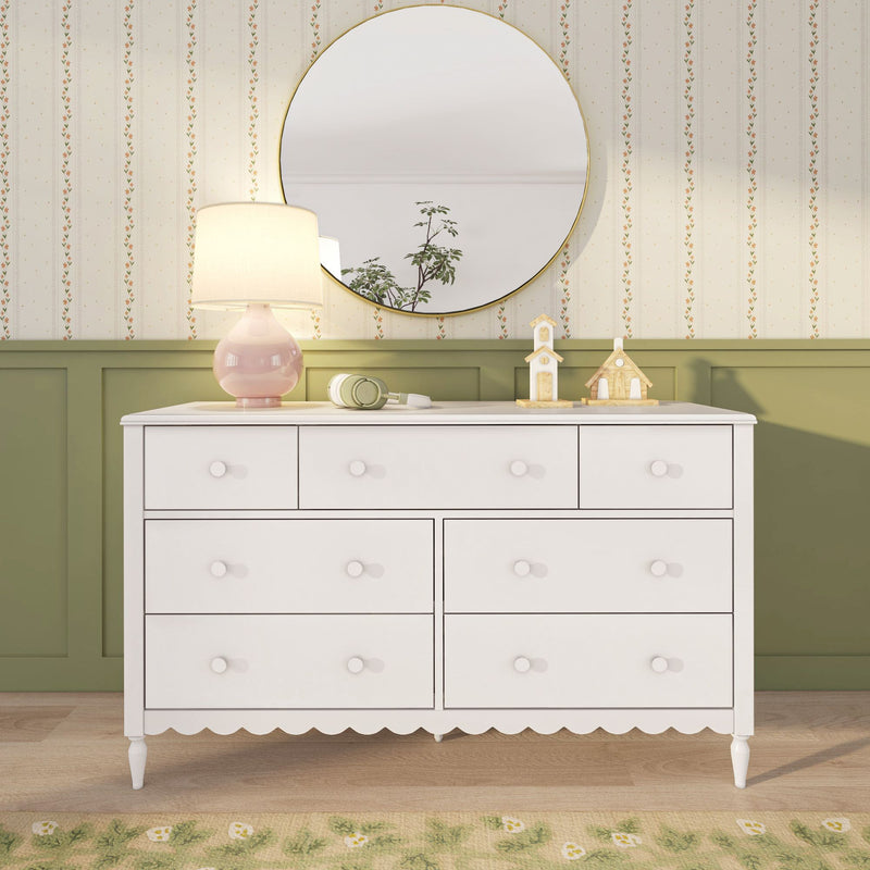 White dresser with round gold-framed mirror, pink table lamp, green headphones and wooden toy houses against vintage floral wallpaper and olive wainscoting — cozy nursery bedroom decor