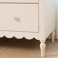 Close-up of cream painted wooden nightstand drawer with round knob, scalloped apron and turned leg on hardwood floor