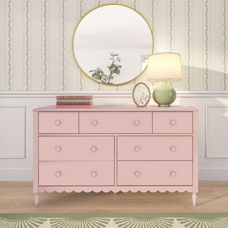 Pink vintage dresser with scalloped trim topped by a round gold mirror, green ceramic table lamp, stacked books and vertical floral wallpaper background