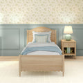 Cozy coastal cottage bedroom with light wood twin bed, pastel blue wainscoting, delicate floral wallpaper, patterned pillows and bedside table lamp