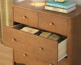 Mid-century modern wooden dresser with brass knobs and warm wood finish, open drawer with neatly folded clothes and a lamp and books on top.