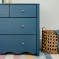 Blue painted wooden dresser with round brass knobs beside woven wicker basket and pastel striped rug, modern nursery storage furniture