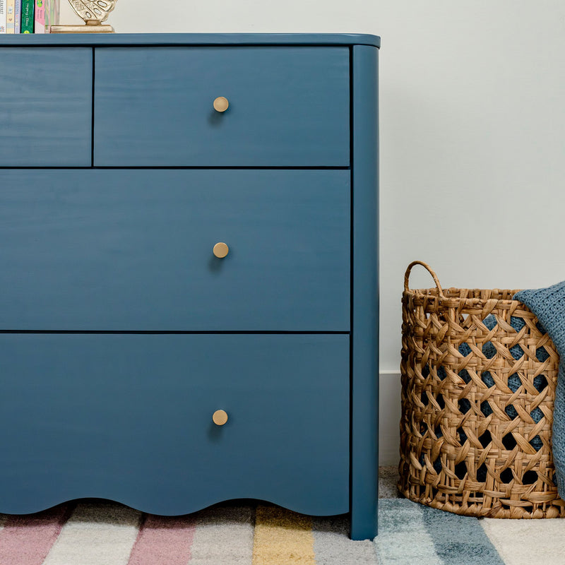 Blue painted wooden dresser with round brass knobs beside woven wicker basket and pastel striped rug, modern nursery storage furniture