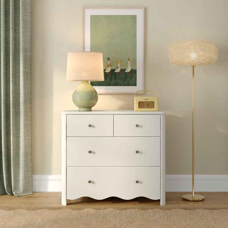 White dresser with green ceramic table lamp, textured gold floor lamp, vintage yellow radio and framed goose print on beige wall with green curtains, modern nursery decor