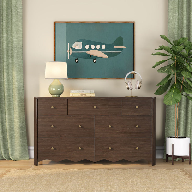 Dark wood dresser with brass knobs beneath framed teal airplane wall art, ceramic table lamp, headphones on a gold stand, potted fiddle‑leaf fig and green curtains