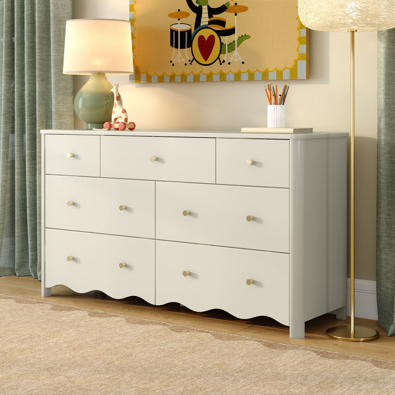 White nursery dresser with round brass knobs and scalloped base, styled with table lamp, wooden giraffe toy, colorful wall art, floor lamp and green curtains on hardwood floor