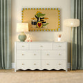 White dresser in kids' nursery with crocodile drummer wall art, ceramic table lamp, wooden giraffe toy, pencil holder and green curtains