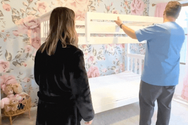 People assembling a white bunk bed frame in a floral-themed bedroom.