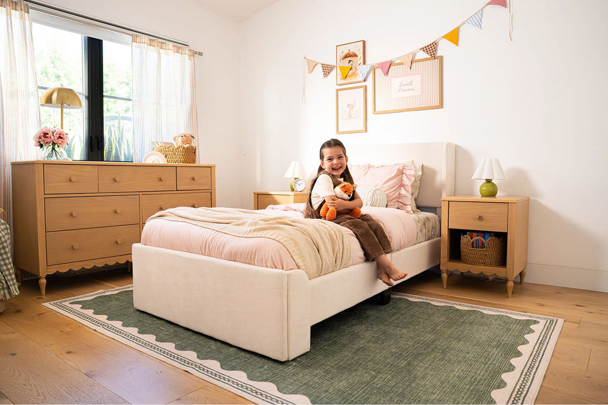 Cozy girl's bedroom decor: child sitting on bed holding stuffed fox, blush pink bedding, wooden dresser and nightstand, bunting wall art, green area rug and natural light.