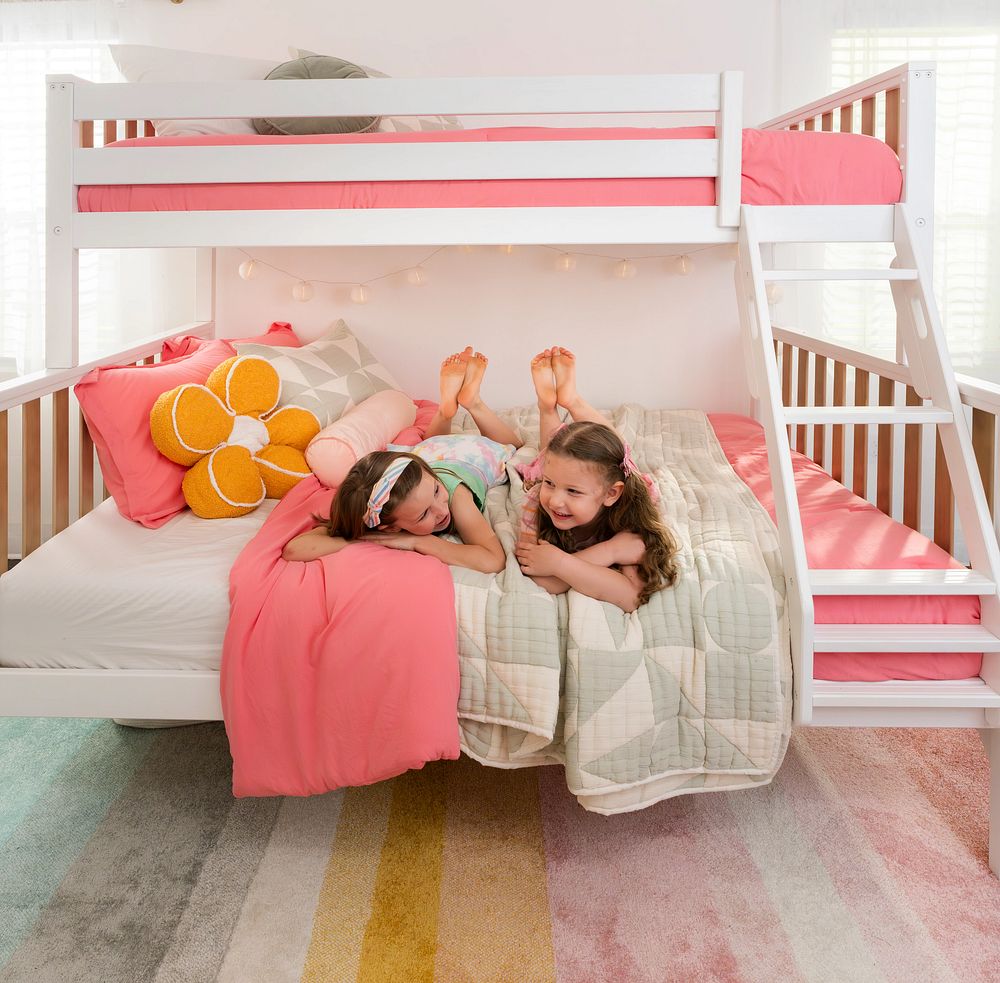 Two young girls lying on the lower bunk of a white wooden bunk bed with pink bedding, a flower-shaped throw pillow, stairs and a colorful striped rug in a bright kids' bedroom.