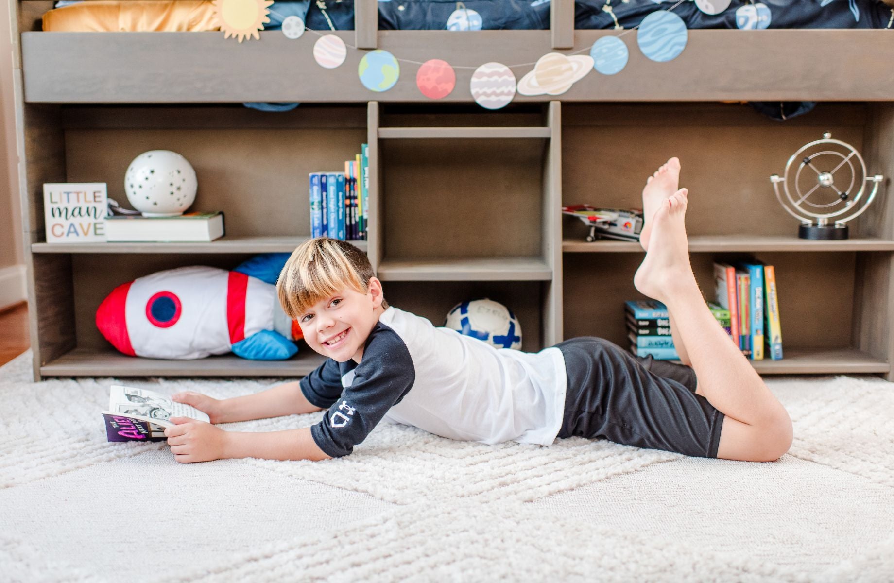 Boy reading a book on a rug in a cozy children's bedroom with space-themed decor.