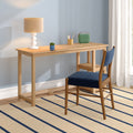 Minimalist home office desk: light wood console with beige table lamp, green portable speaker, pencil holder and potted succulent, navy upholstered chair, blue wall and striped rug