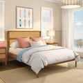 Sunlit coastal bedroom with rattan headboard, white bedding, coral accent pillows and blue throw, framed surfboard art, bedside lamp and large ocean-view window.