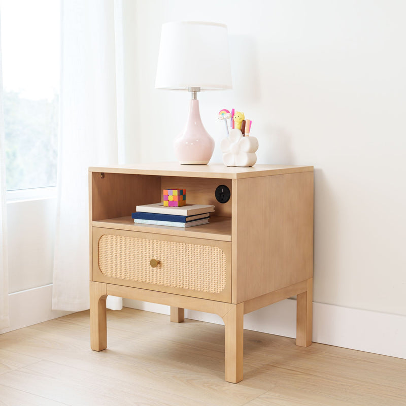 Light wood bedside table nightstand with rattan drawer, pink ceramic table lamp with white shade, flower-shaped pen holder with colorful pens, stacked books and a small toy cube in a bright minimalist nursery/kids room decor