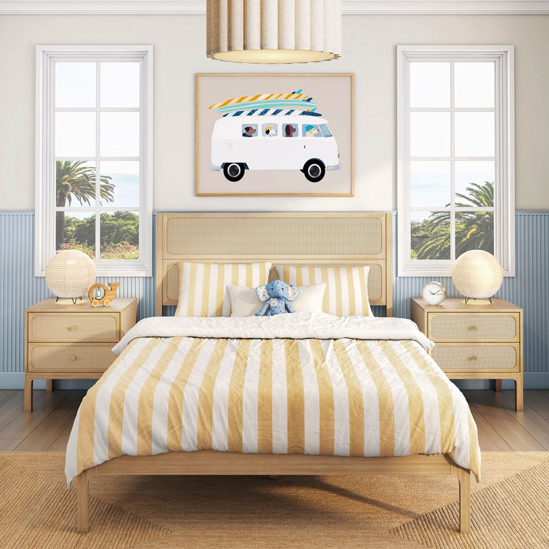 Beach-themed kids bedroom with yellow-and-white striped bedding, rattan headboard and light wood bed, matching nightstands with round lamps, surf-van wall art and twin windows overlooking palm trees and ocean.
