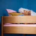 Stuffed teddy bear on wooden bunk bed with pink bedding and colorful geometric pillows against a blue bedroom wall