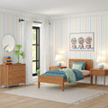 Children's bedroom with mid-century wooden twin bed and blue bedding, pastel striped wallpaper, colorful gumball machine wall art, matching nightstands with lamps, wooden dresser and mirror, houseplant by a sunlit window, light hardwood floor and neutral area rug