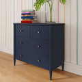 Navy blue mid-century modern dresser with brass knobs topped by colorful stacked books and a glass vase with monstera leaves, against pastel striped wallpaper on a hardwood floor.