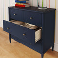 Navy blue mid-century modern dresser with open drawer showing folded clothes, brass knobs, stacked books and glass vase on hardwood floor against pastel striped wallpaper