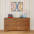 Mid-century modern six-drawer wooden dresser on hardwood floor against pastel-striped wallpaper, topped with a blue globe table lamp, teal alarm clock, colorful stacked books and framed rainbow abstract art
