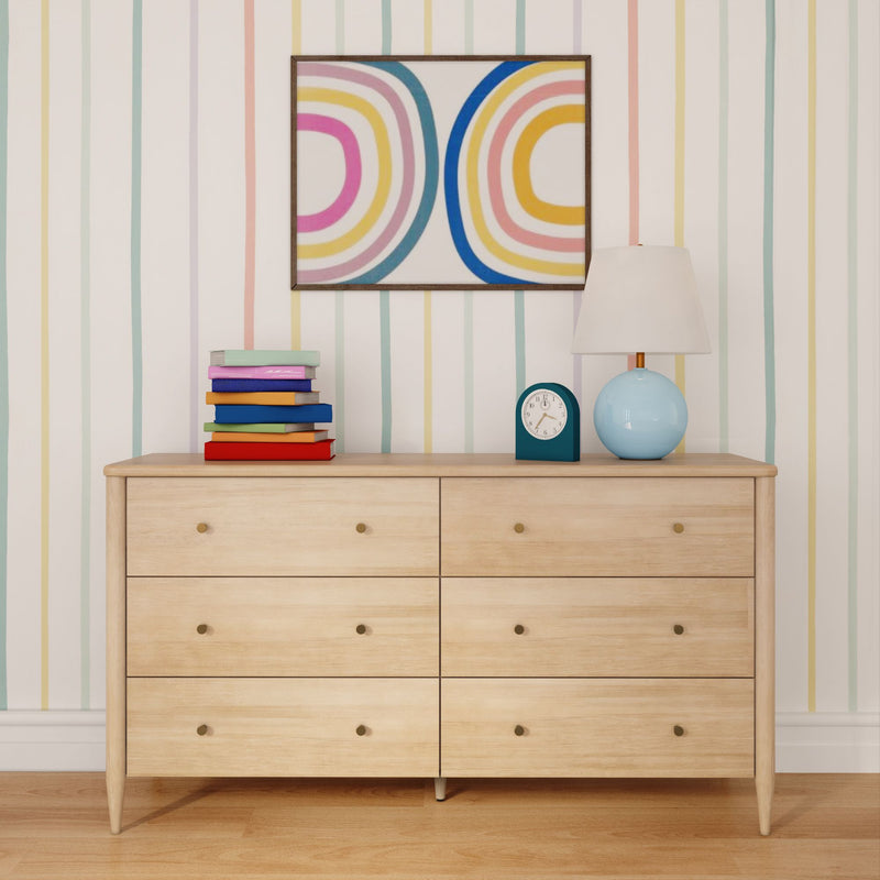 Light wood mid-century dresser with six drawers, pastel striped wallpaper, stack of colorful books, blue table lamp, teal alarm clock and abstract rainbow wall art in a nursery
