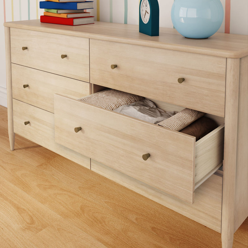 Light wood dresser with an open drawer showing folded bedding and pillows, stacked books, a clock and blue vase on top, hardwood floor