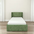 Green upholstered twin bed with white mattress in a modern minimalist bedroom featuring hardwood floors, neutral walls, and sheer white curtains