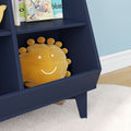 Navy blue kids bookshelf in a modern nursery holding a smiling yellow sun plush pillow and illustrated children's books, set against a light blue wall with white shag rug and hardwood floor.