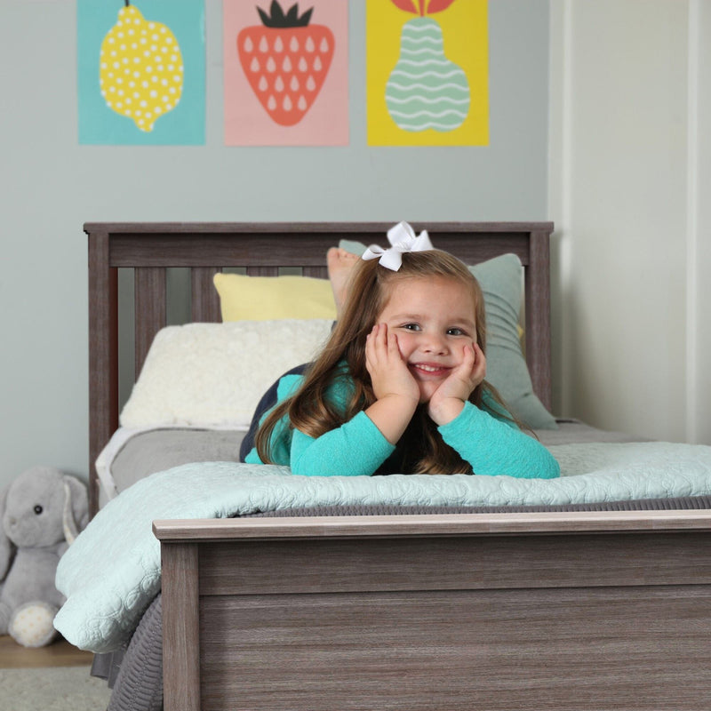Smiling young girl lying on bed in a pastel kids' bedroom with colorful fruit wall art, white bow and stuffed elephant